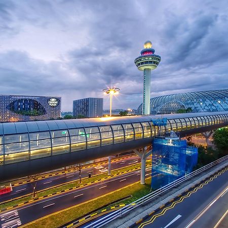 فندق كراون بلازا مطار شانجي سنغافورة المظهر الخارجي الصورة The photo depicts an airport scene, likely featuring a modern terminal structure with a distinctive, futuristic design. In the foreground, there is a pedestrian bridge or walkway, along with roads leading to the terminal. A control tower can be seen 