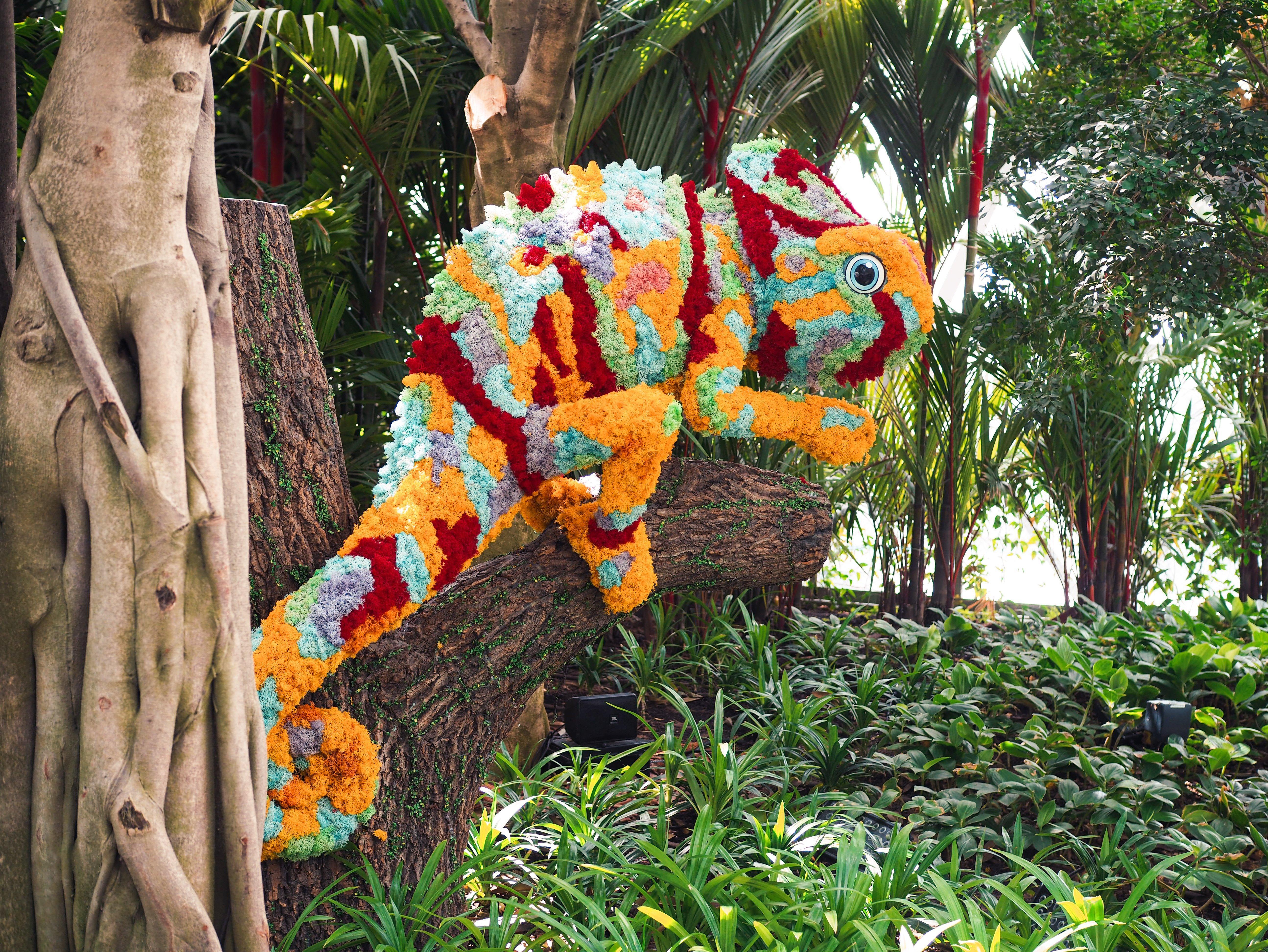 فندق كراون بلازا مطار شانجي سنغافورة المظهر الخارجي الصورة The photo shows a vibrant and colorful chameleon sculpture made from flowers and plant materials. The chameleon is positioned on a tree trunk, blending in with the lush greenery of its surroundings. The colors of the sculpture include shades of orang