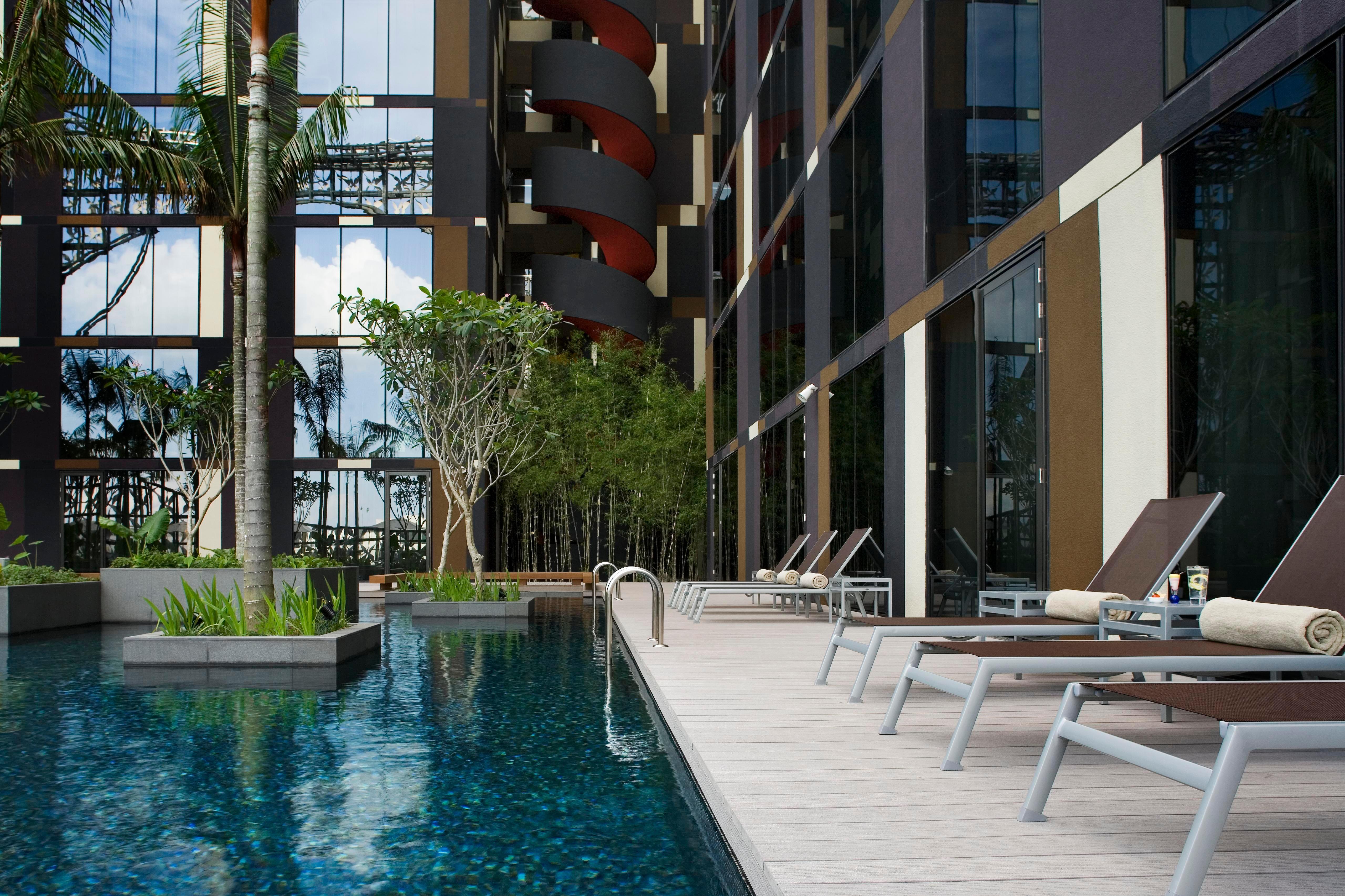 فندق كراون بلازا مطار شانجي سنغافورة المرافق الصورة The photo shows a modern swimming pool area adjacent to a building. The pool is surrounded by sleek wooden decking and features a clear blue water surface. There are several lounge chairs positioned around the pool for relaxation. Lush greenery, incl