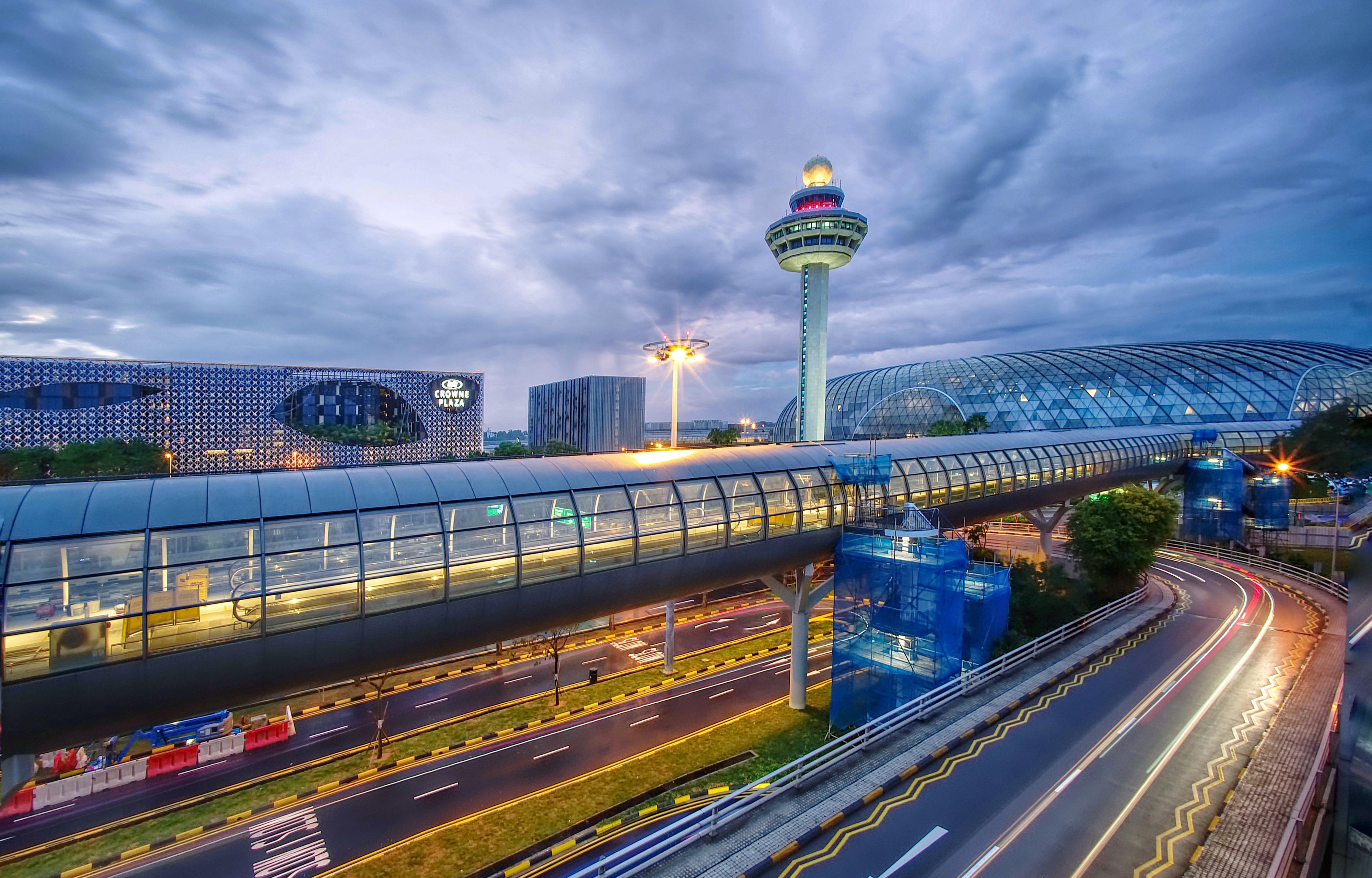 فندق كراون بلازا مطار شانجي سنغافورة المظهر الخارجي الصورة The photo depicts an airport scene, likely featuring a modern terminal structure with a distinctive, futuristic design. In the foreground, there is a pedestrian bridge or walkway, along with roads leading to the terminal. A control tower can be seen 
