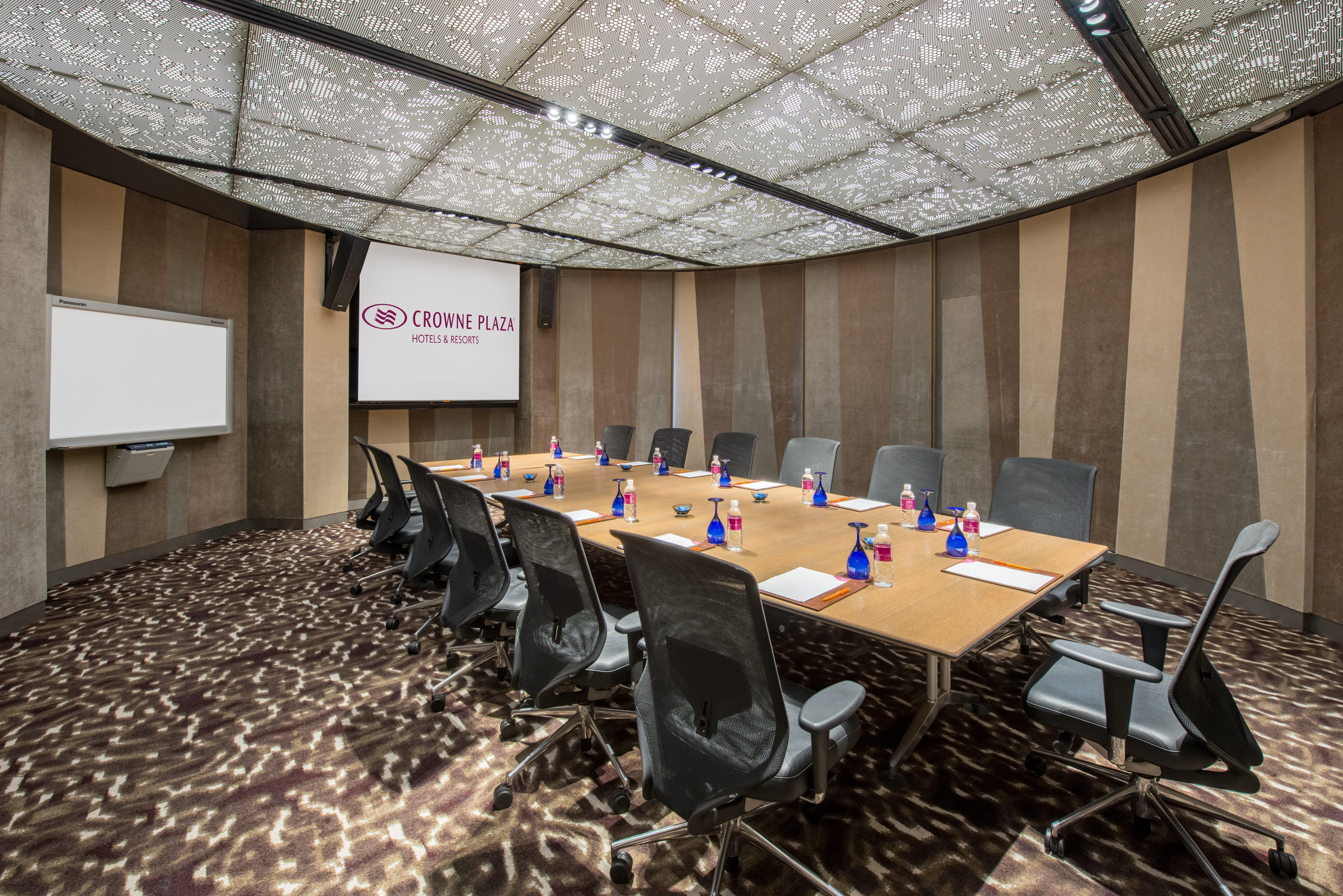 فندق كراون بلازا مطار شانجي سنغافورة المظهر الخارجي الصورة The photo shows a modern conference room. It features a large, rectangular wooden table surrounded by ergonomic black office chairs. On the table, there are small blue vases, each containing flowers. The room has a unique, patterned ceiling that enha