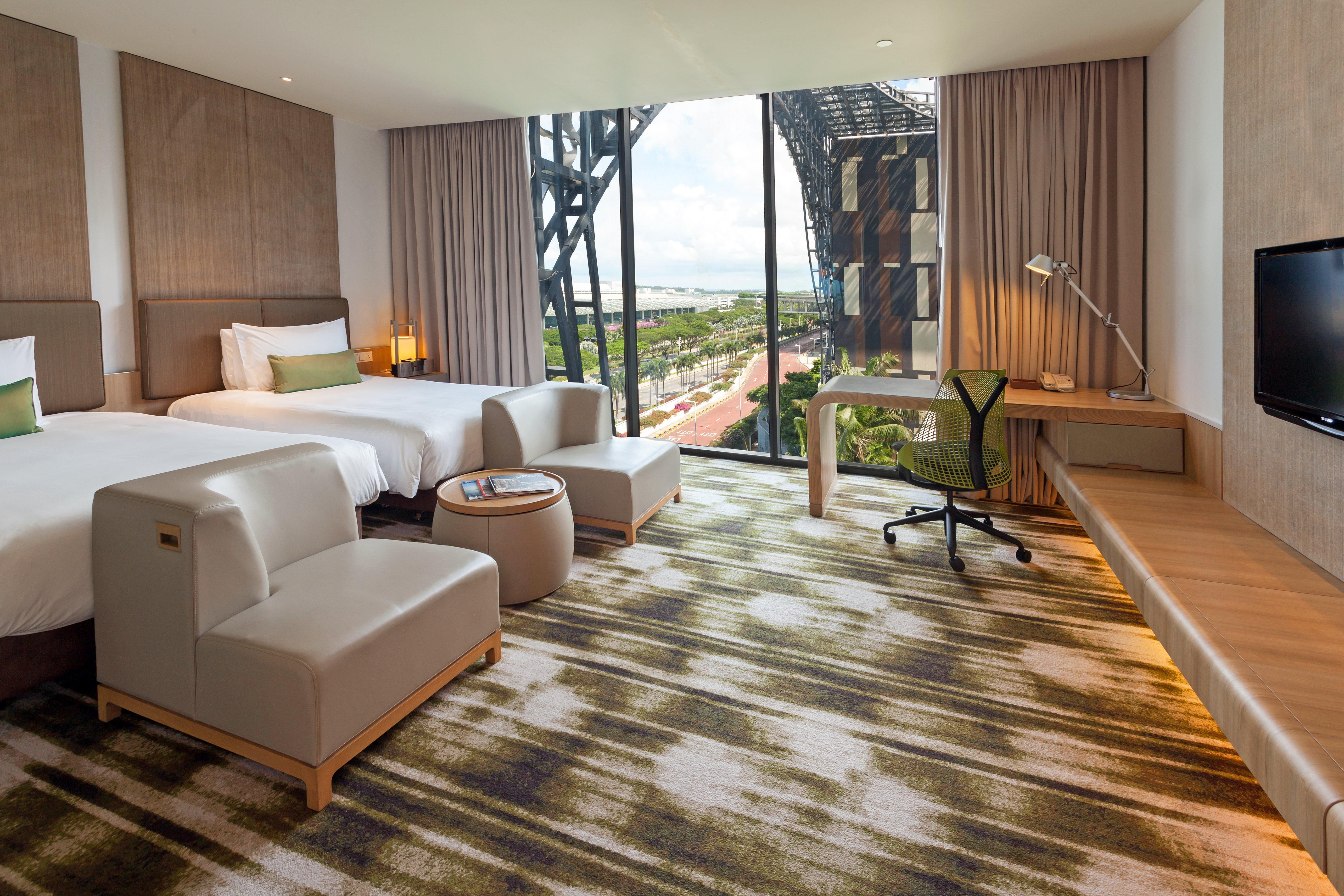 فندق كراون بلازا مطار شانجي سنغافورة المظهر الخارجي الصورة The photo depicts a modern hotel room featuring two beds with white linens and a clean, minimalist design. There are two stylish armchairs and a small round table, creating a cozy seating area. The room includes a large window that offers a view of t