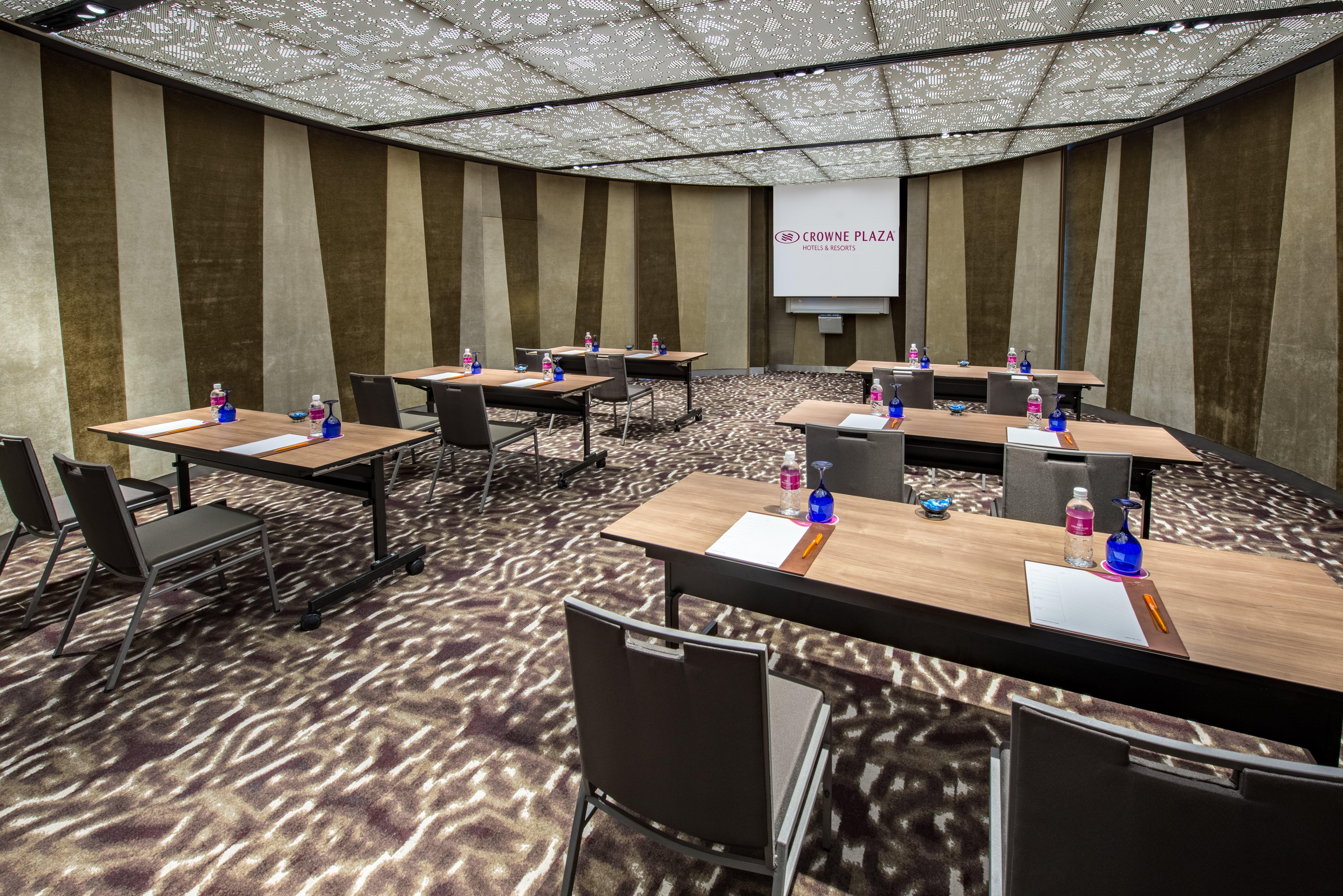 فندق كراون بلازا مطار شانجي سنغافورة المظهر الخارجي الصورة The photo shows a modern conference room set up for a meeting or presentation. There are several wooden tables arranged in a U-shape, each equipped with a notepad and pen. Bottled water is placed in blue containers on the tables. The room features a 
