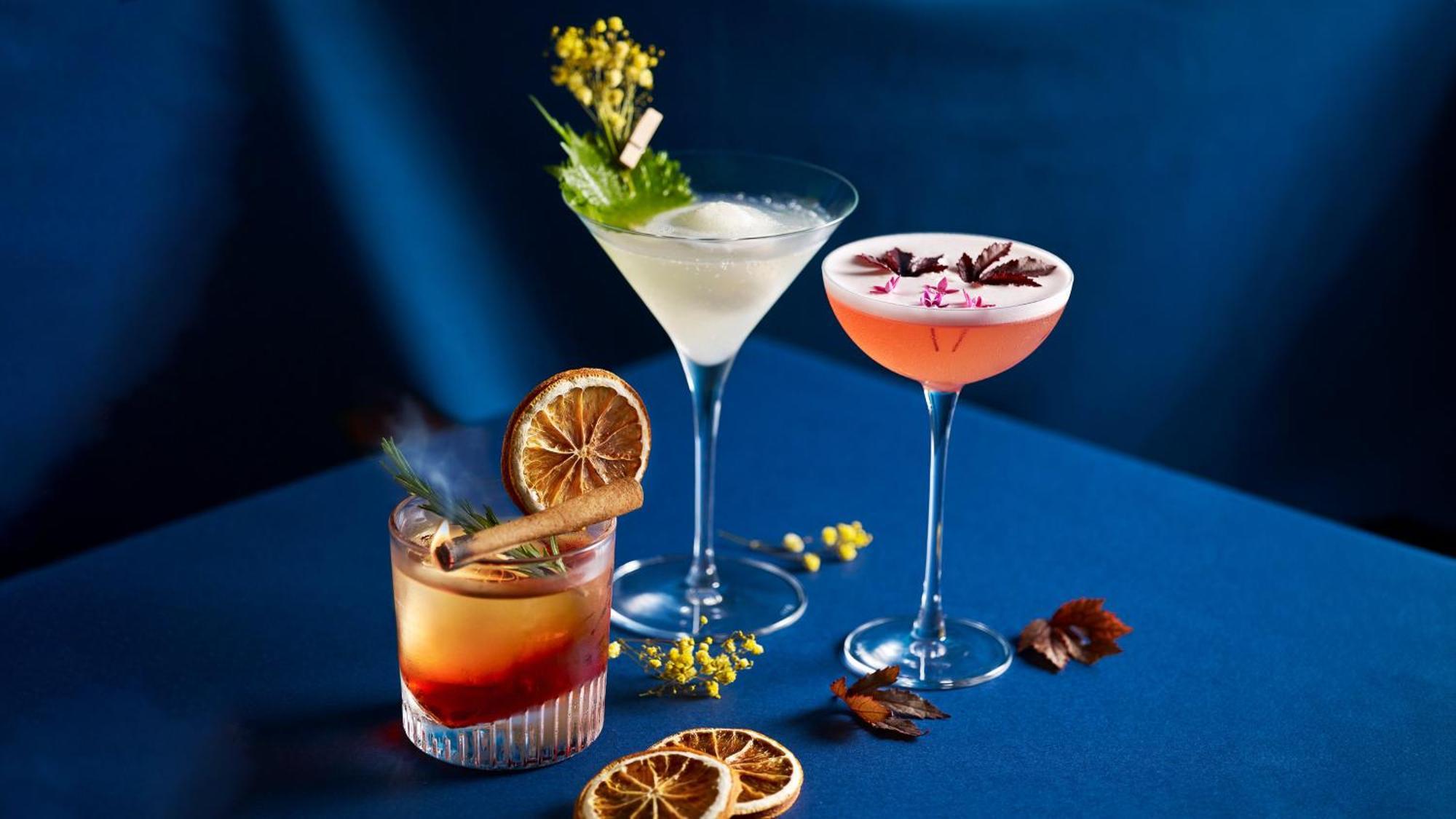 فندق كراون بلازا مطار شانجي سنغافورة المظهر الخارجي الصورة The photo shows a stylish arrangement of three cocktails on a blue surface. 

1. **Left Cocktail**: A lowball glass containing a drink with a reddish hue, garnished with a dried orange slice and a cinnamon stick. 

2. **Middle Cocktail**: A classic m