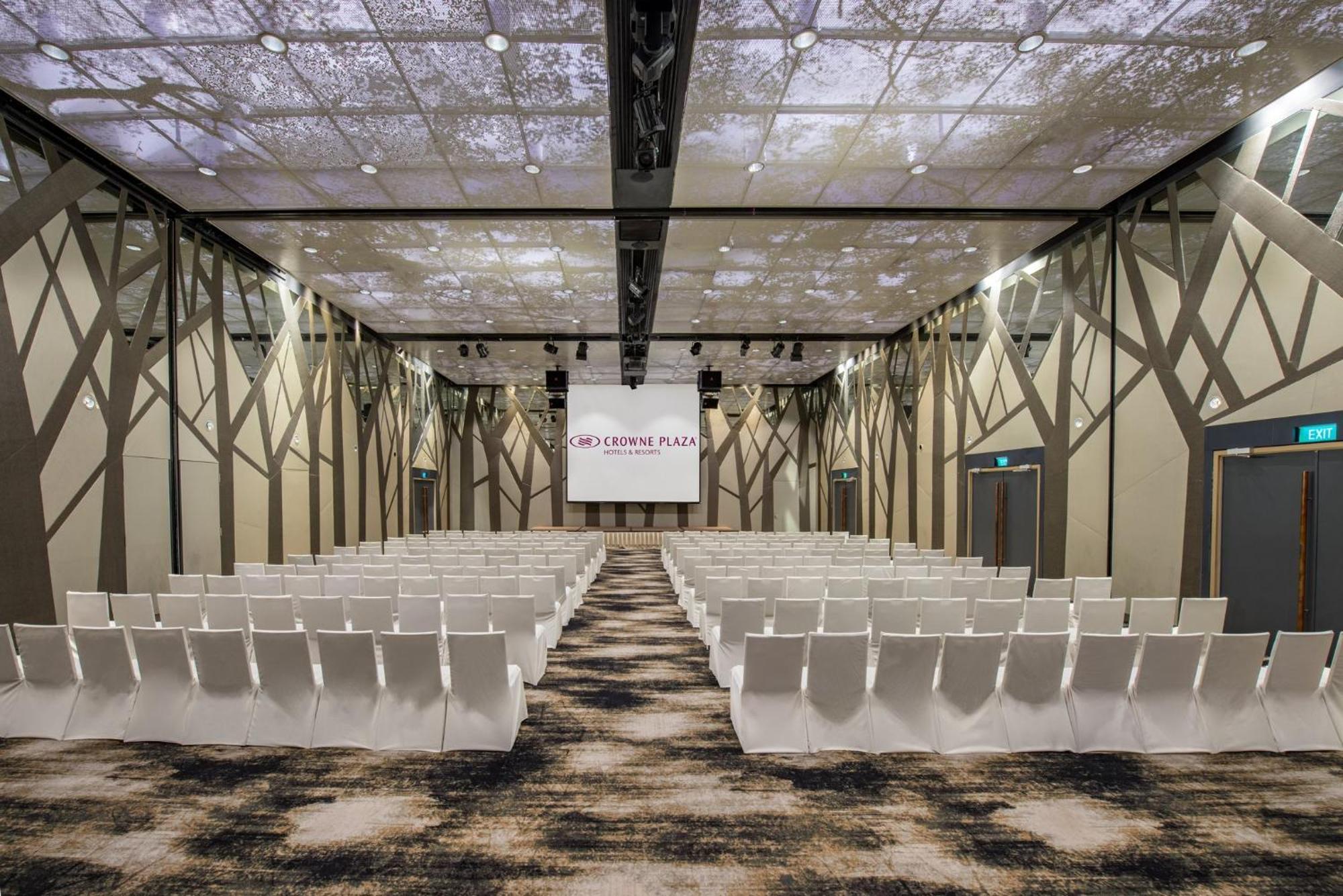 فندق كراون بلازا مطار شانجي سنغافورة المظهر الخارجي الصورة The photo shows a modern conference or event space. The room features rows of white chairs arranged in a theater-style layout, facing a large screen with a simple logo. The ceiling is designed to resemble a natural setting, with images that suggest t