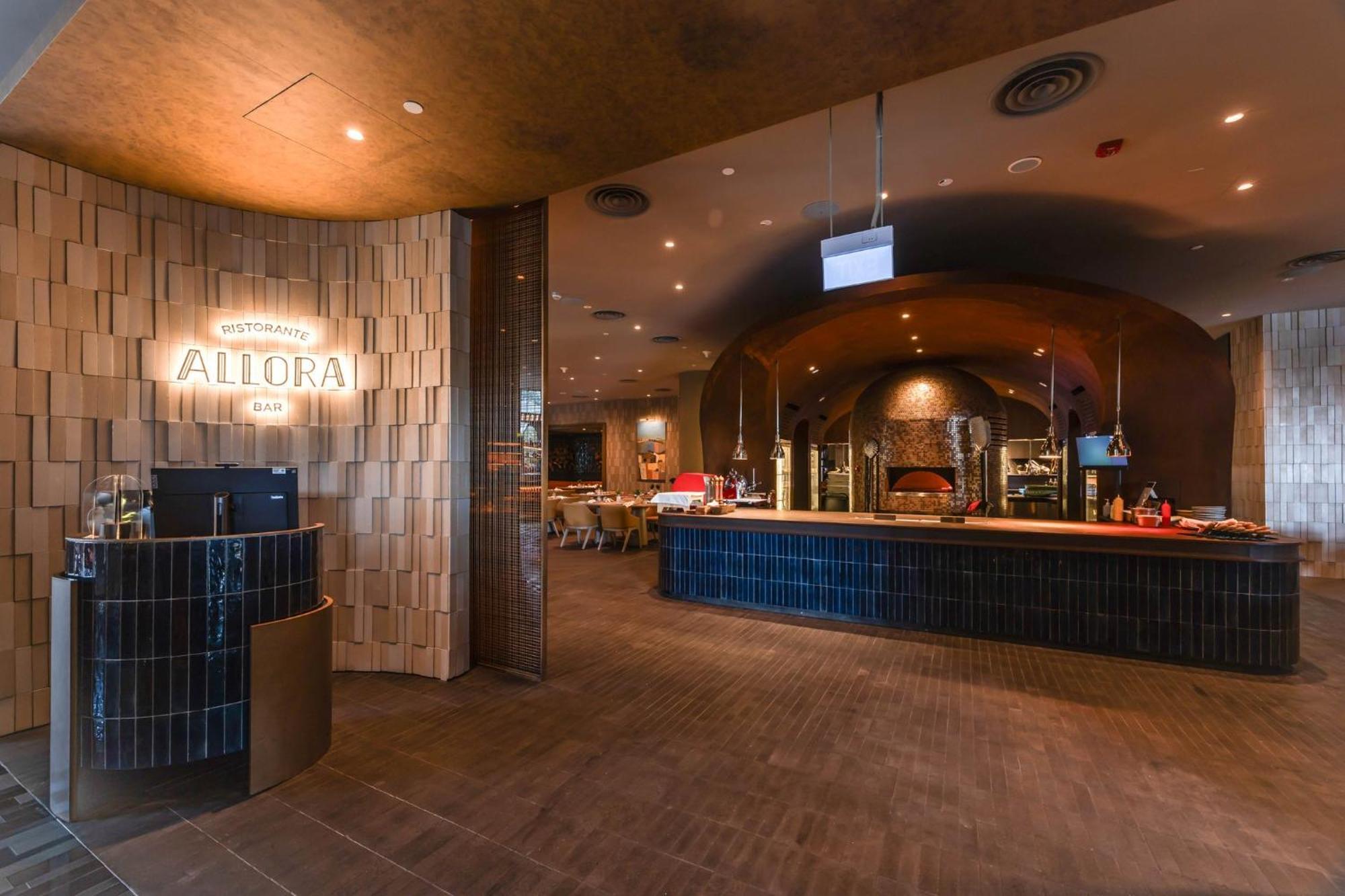 فندق كراون بلازا مطار شانجي سنغافورة المظهر الخارجي الصورة The photo shows the interior of a restaurant named "Allora." The design features a contemporary style with a warm color palette. In the foreground, there is a curved bar area with a sign indicating the restaurant's name. The walls are adorned with te