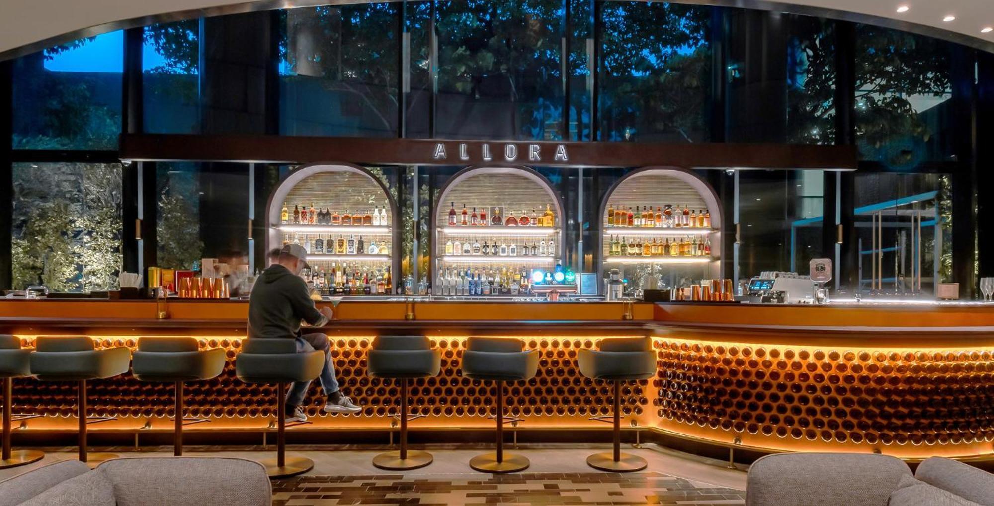 فندق كراون بلازا مطار شانجي سنغافورة المظهر الخارجي الصورة The photo depicts a modern bar interior with a sleek design. The bar is illuminated with warm lighting, highlighting a wide selection of alcohol displayed on shelves behind the counter. There are several high-backed stools at the bar, and a person is