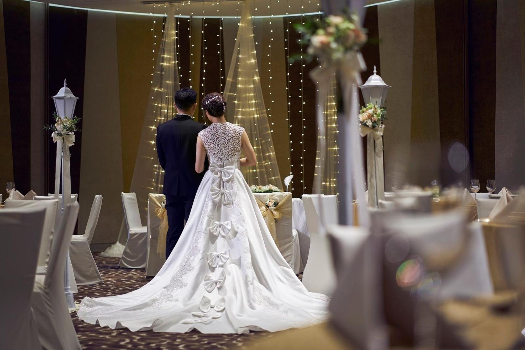 فندق كراون بلازا مطار شانجي سنغافورة المظهر الخارجي الصورة The photo features a couple at a wedding venue. The bride, wearing an elegant white gown with intricate details and a long train, is seen from behind. The groom stands beside her, dressed in a dark suit. The background showcases a beautifully decorat