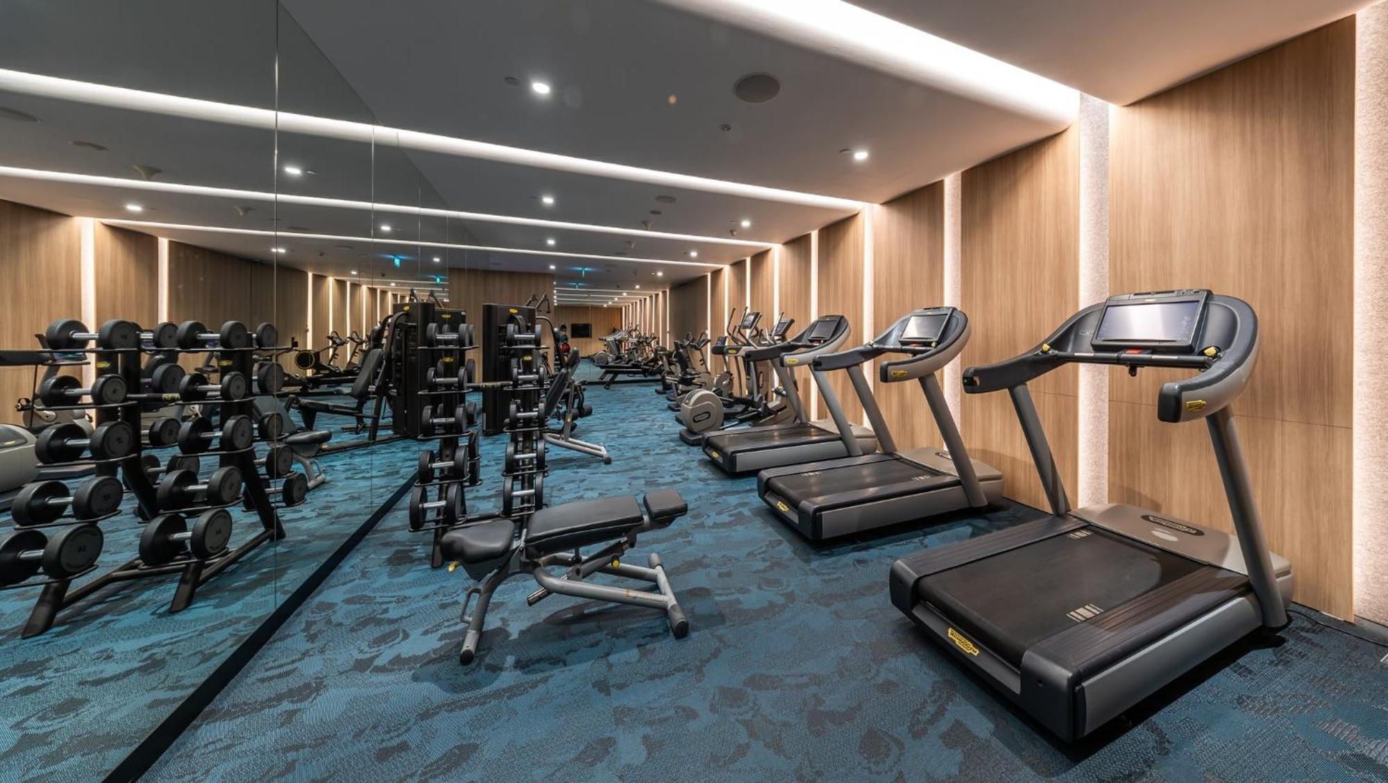 فندق كراون بلازا مطار شانجي سنغافورة المظهر الخارجي الصورة The photo shows a well-equipped gym or fitness room. On one side, there are several weightlifting benches along with dumbbells arranged on racks. On the other side, there are multiple treadmills lined up, suggesting a space designed for both strength
