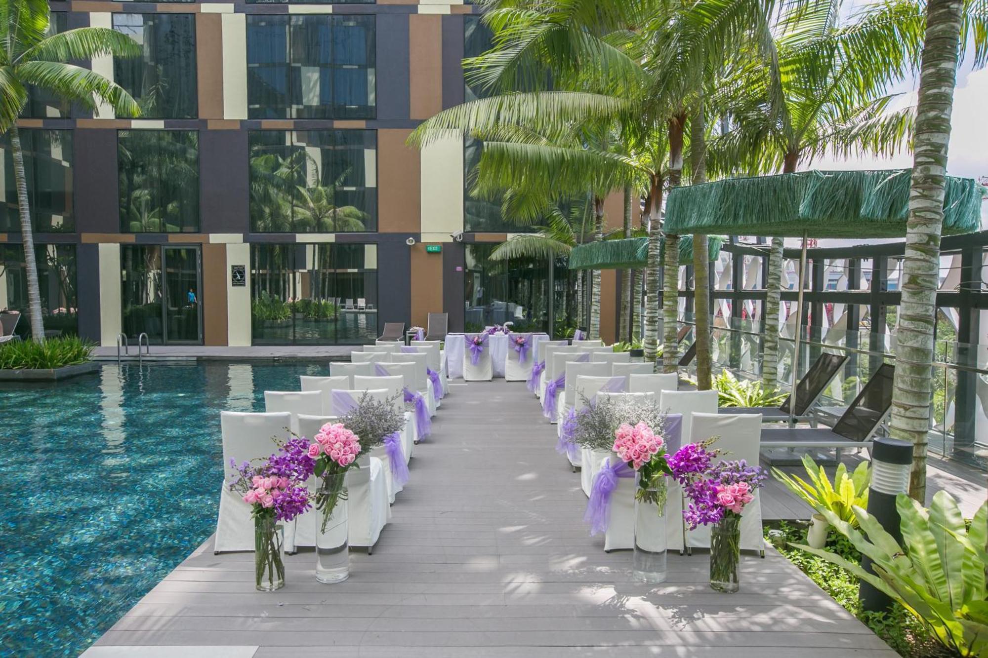 فندق كراون بلازا مطار شانجي سنغافورة المظهر الخارجي الصورة The photo shows a beautifully arranged outdoor setting for a wedding or a similar event. There is a long aisle made of wooden planks leading to a pool area, flanked by neatly arranged chairs. Each chair is adorned with purple ribbons and decorated wi