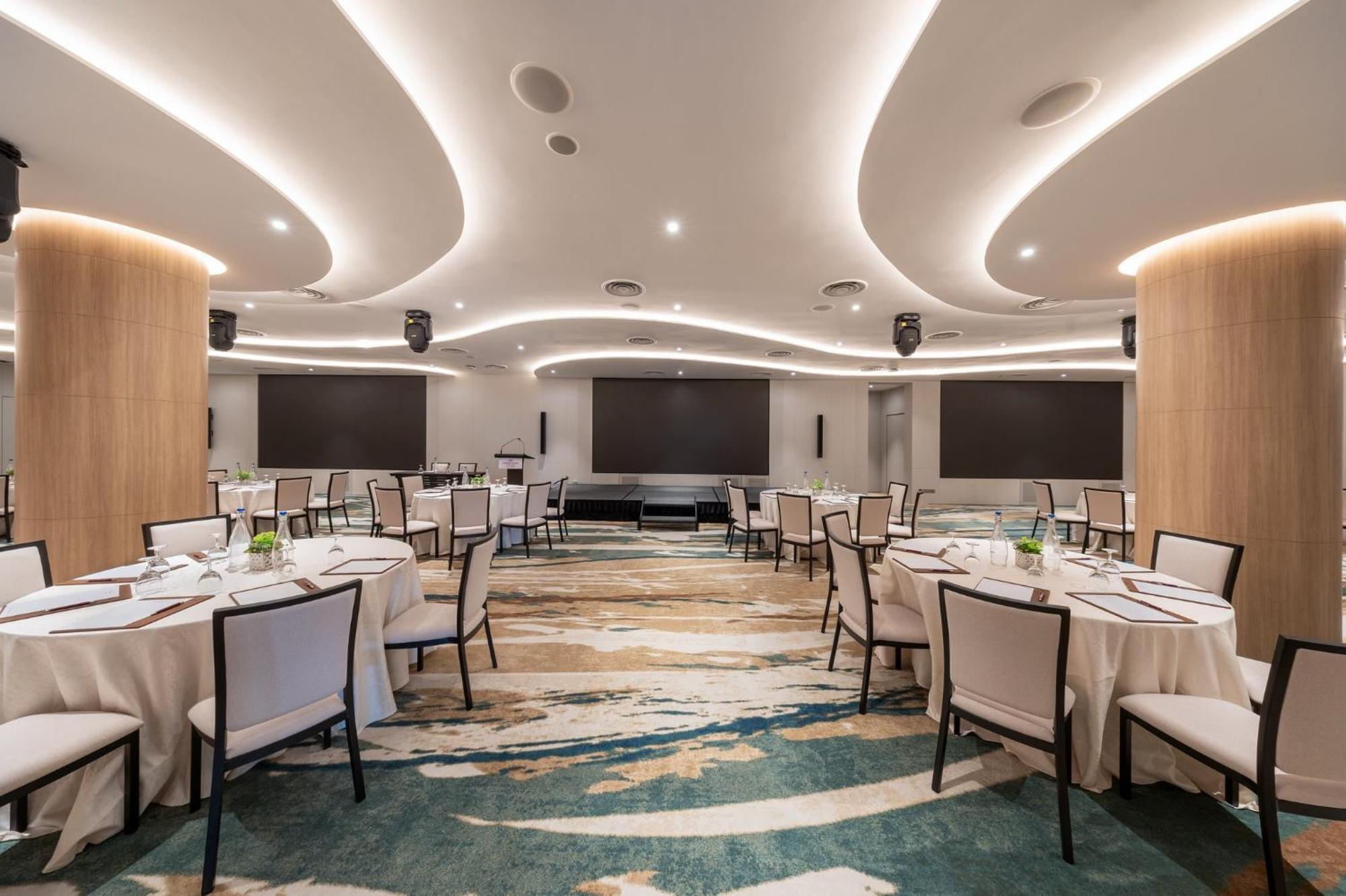 فندق كراون بلازا مطار شانجي سنغافورة المظهر الخارجي الصورة The photo shows a modern banquet or conference room. The room features elegantly arranged round tables with white tablecloths and chairs. The flooring has an artistic design that combines various shades of blue and beige. The ceiling is accentuated w