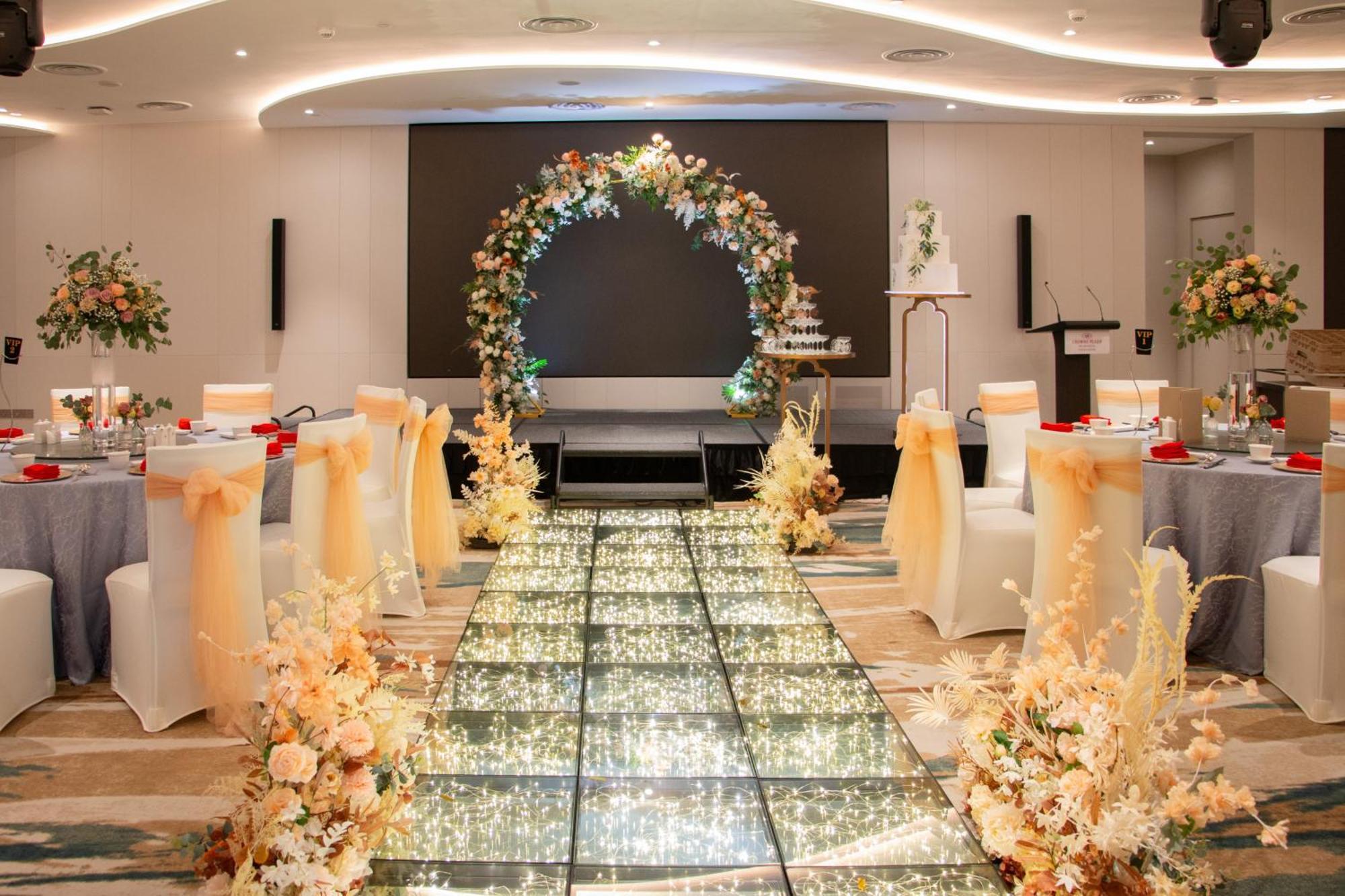 فندق كراون بلازا مطار شانجي سنغافورة المظهر الخارجي الصورة The image depicts an elegantly decorated event space, likely for a wedding or formal occasion. In the center, there is a flower-adorned archway in warm colors, creating a focal point. A sparkling, illuminated pathway leads towards the arch, bordered 