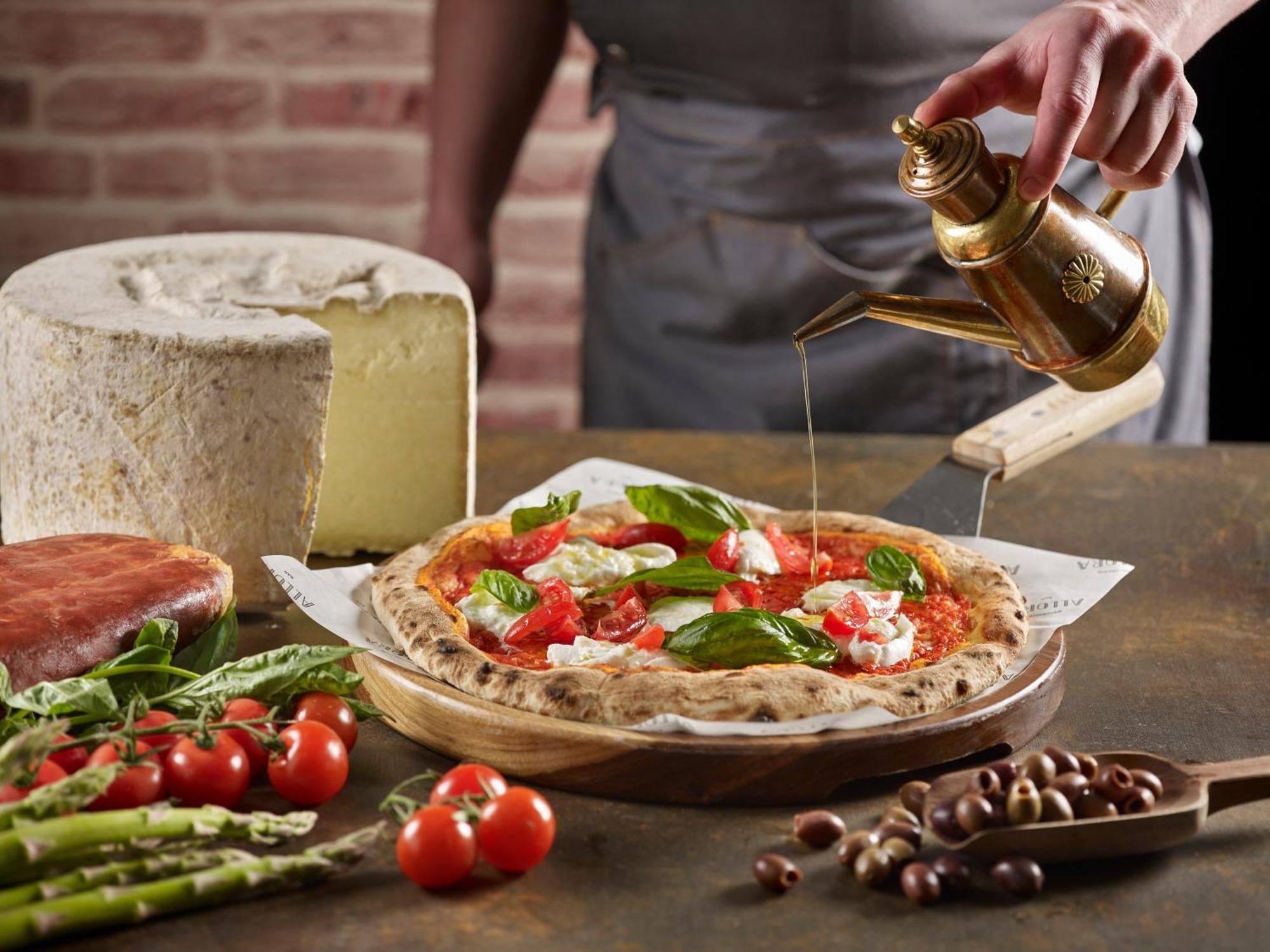 فندق كراون بلازا مطار شانجي سنغافورة المظهر الخارجي الصورة The photo features a freshly made pizza being drizzled with oil. The pizza is topped with ingredients such as fresh basil and tomatoes, and has a golden, crispy crust. In the background, there is a block of cheese and a piece of ham. Surrounding the 