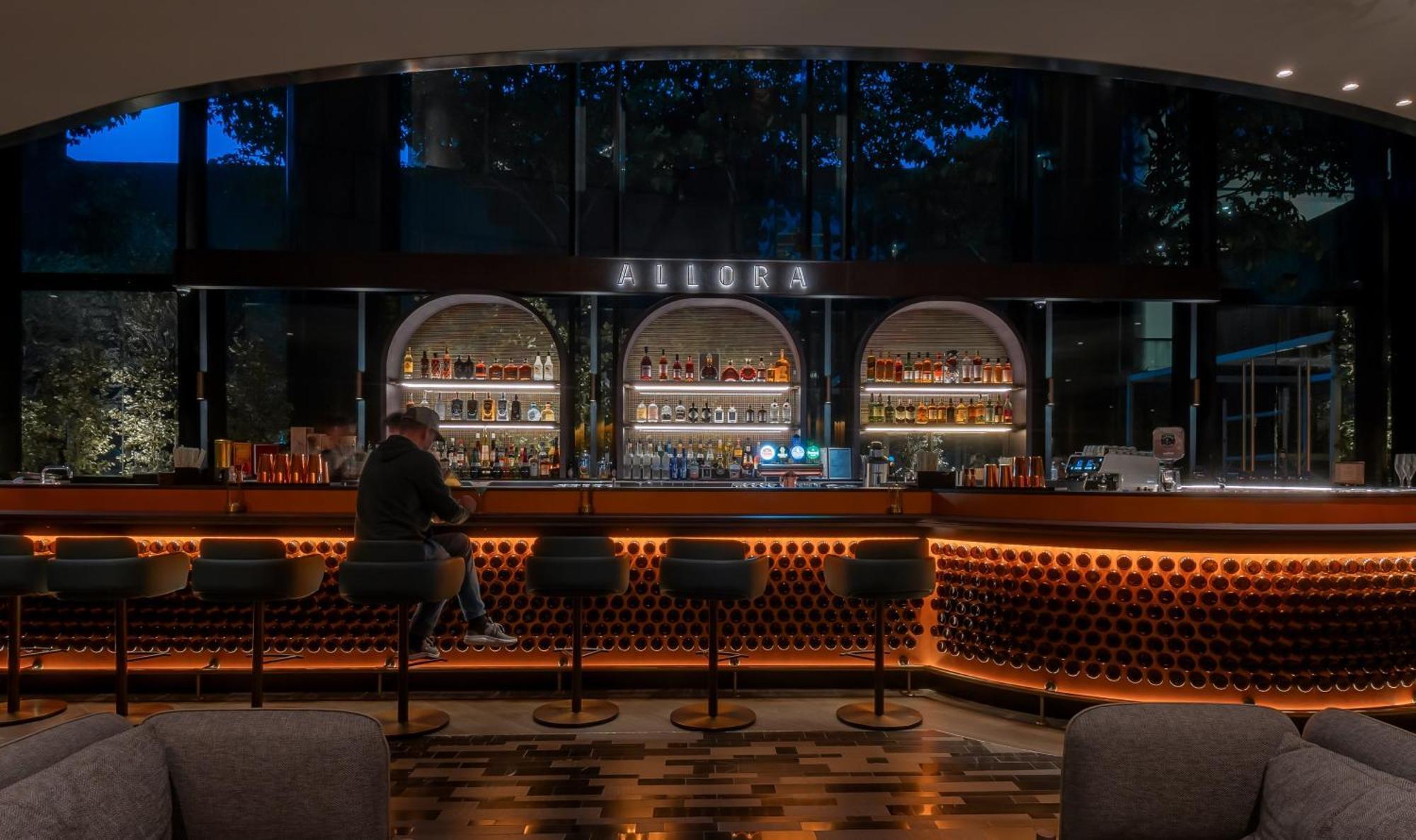 فندق كراون بلازا مطار شانجي سنغافورة المظهر الخارجي الصورة The photo shows a bar area with a modern and stylish design. In the center, there is a well-lit bar counter featuring a variety of alcoholic beverages displayed on shelves behind it. The bar has a sleek, contemporary look with circular light fixtures