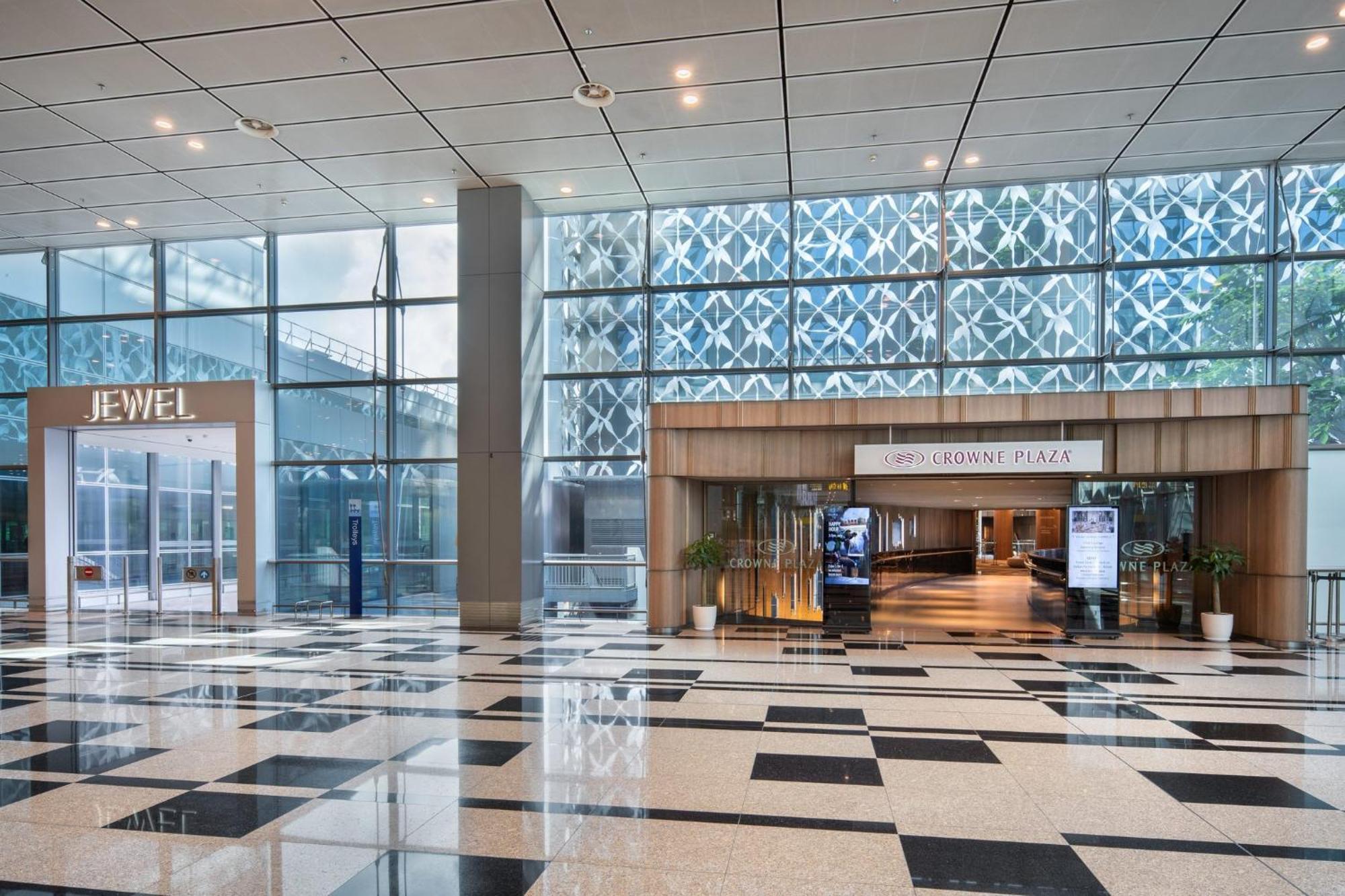 فندق كراون بلازا مطار شانجي سنغافورة المظهر الخارجي الصورة The photo shows an indoor area with a modern architectural design featuring large glass windows that allow natural light to enter. On the left side, there is a sign for "JEWEL," indicating a retail or entertainment space. In the center, there is a wo