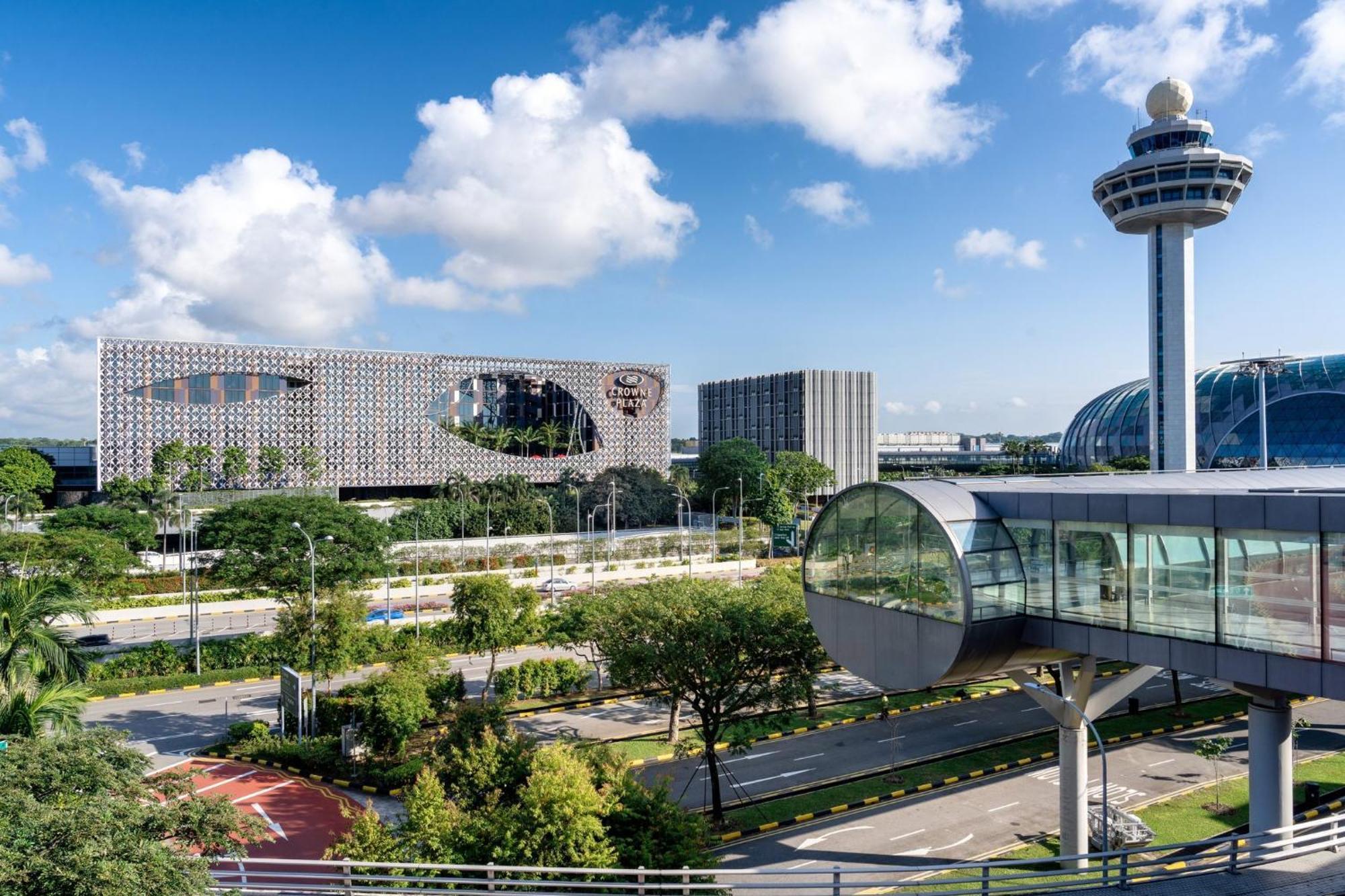 فندق كراون بلازا مطار شانجي سنغافورة المظهر الخارجي الصورة The photo shows a modern urban landscape with a clear blue sky and fluffy white clouds. In the foreground, there is a landscaped area with greenery and a road. Prominently featured is an interesting architectural structure, likely a building with a u