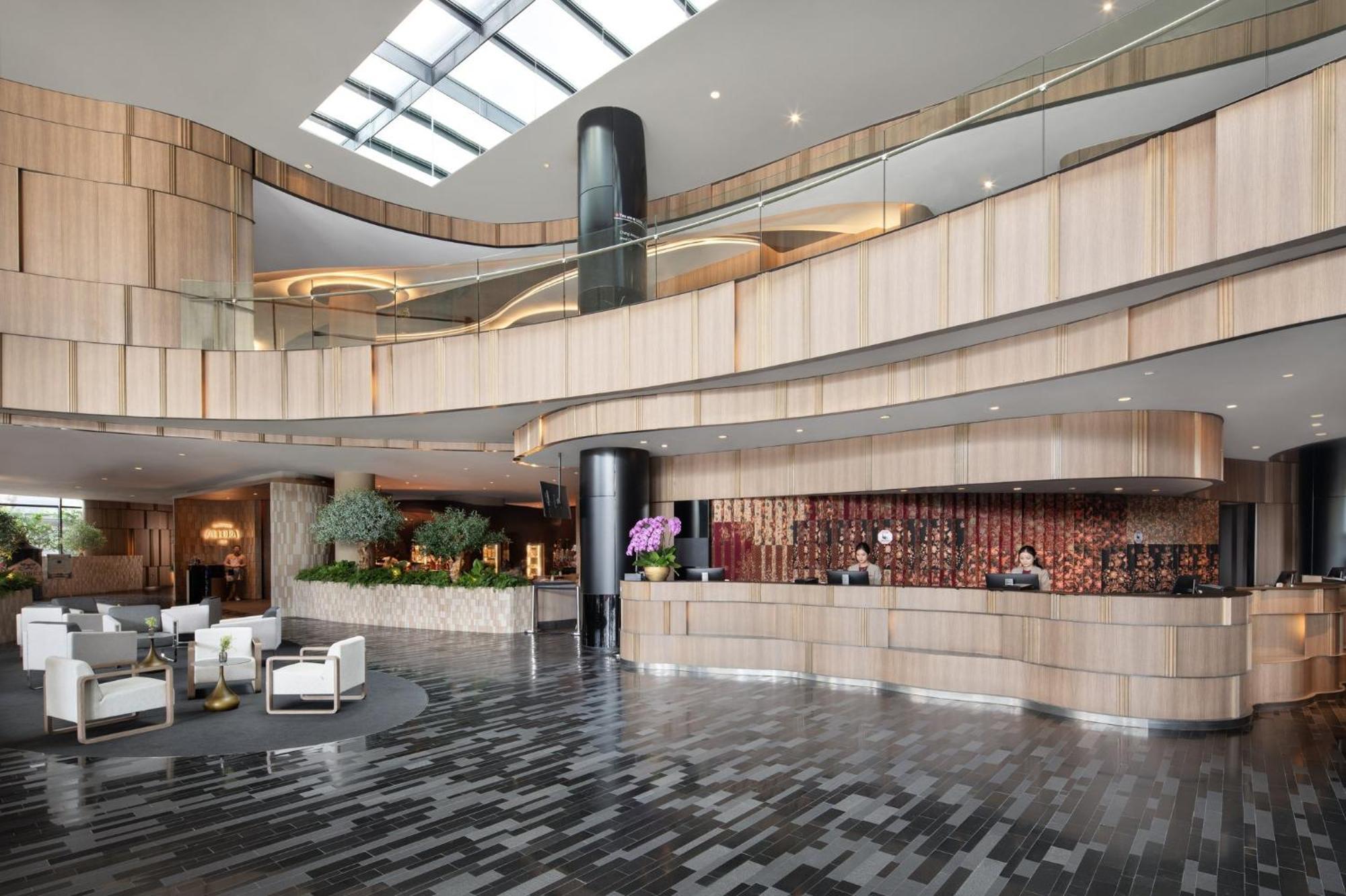 فندق كراون بلازا مطار شانجي سنغافورة المظهر الخارجي الصورة The photo shows a modern hotel lobby with a sleek and stylish design. The reception area features a curved wooden counter, with staff members attending to guests. The interior design includes a mix of wood and stone materials, creating a warm and inv