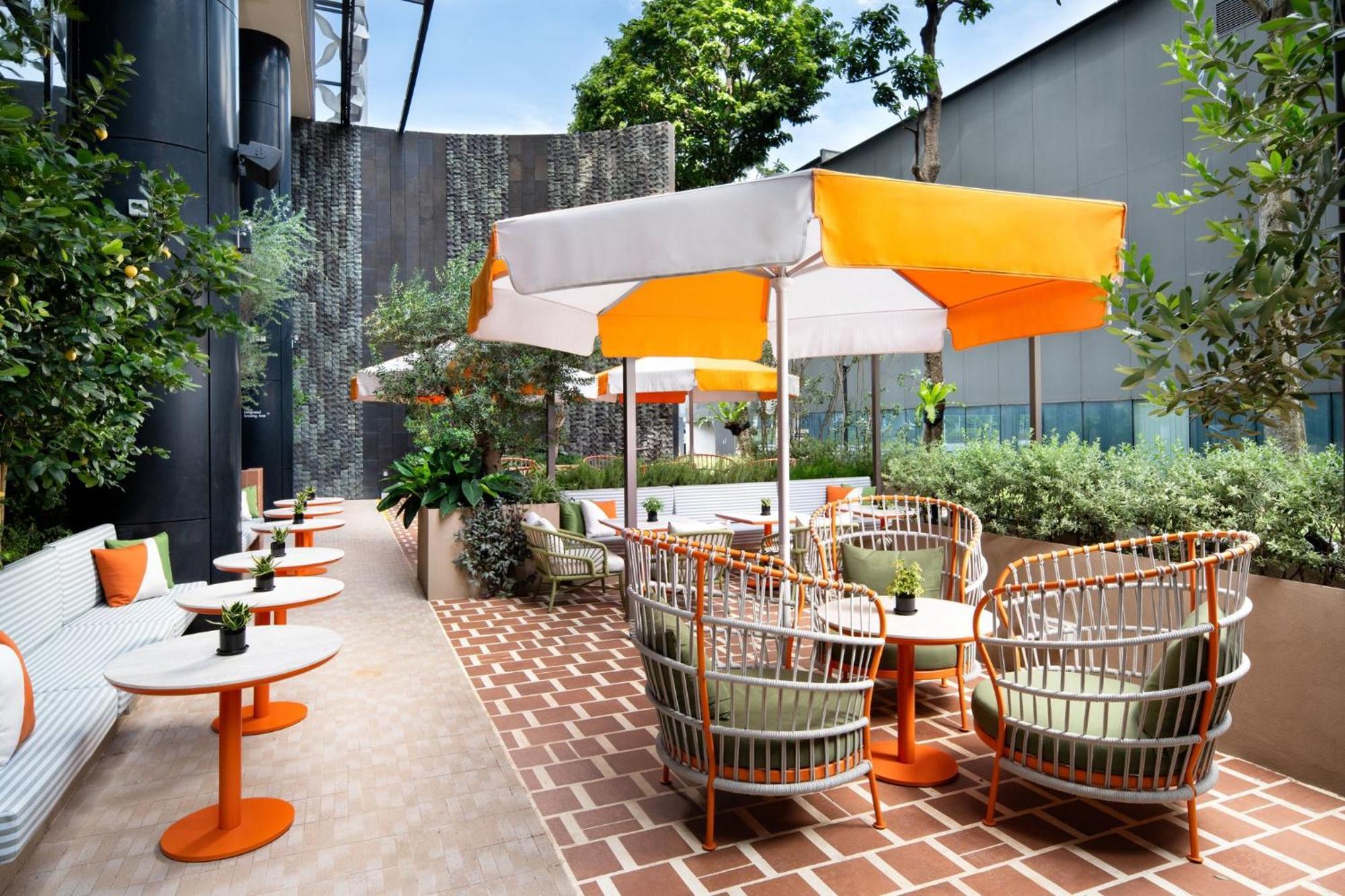 فندق كراون بلازا مطار شانجي سنغافورة المظهر الخارجي الصورة The photo depicts an outdoor seating area featuring several tables and chairs. The tables have orange bases and white tops, while the seating comprises rattan-style chairs with cushions. Each table is accompanied by a large umbrella with a white and 
