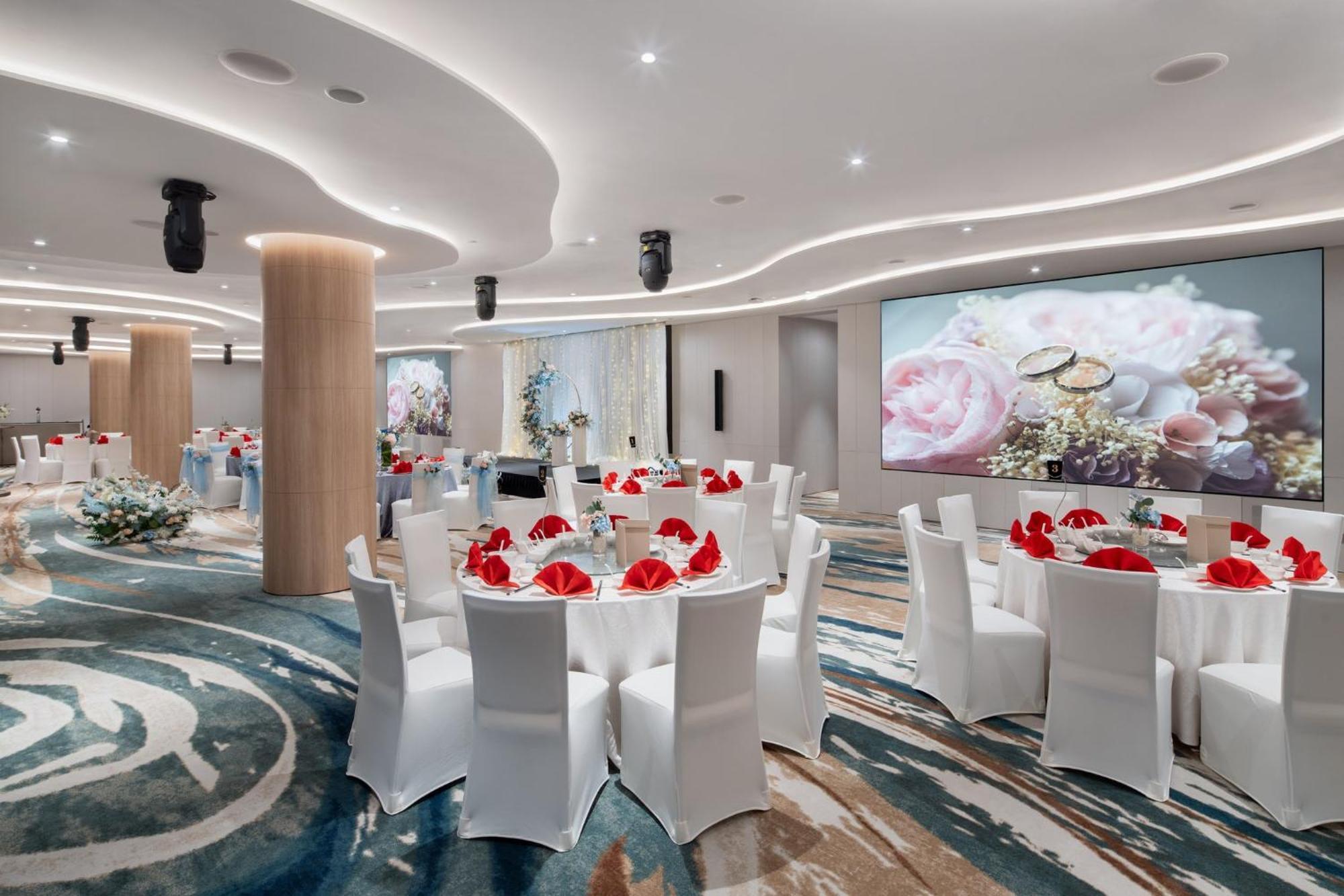 فندق كراون بلازا مطار شانجي سنغافورة المظهر الخارجي الصورة The photo depicts an elegantly designed banquet or event hall. It features round dining tables covered with white tablecloths, each adorned with red napkins. The chairs are also white, creating a cohesive color scheme. The room has modern lighting an