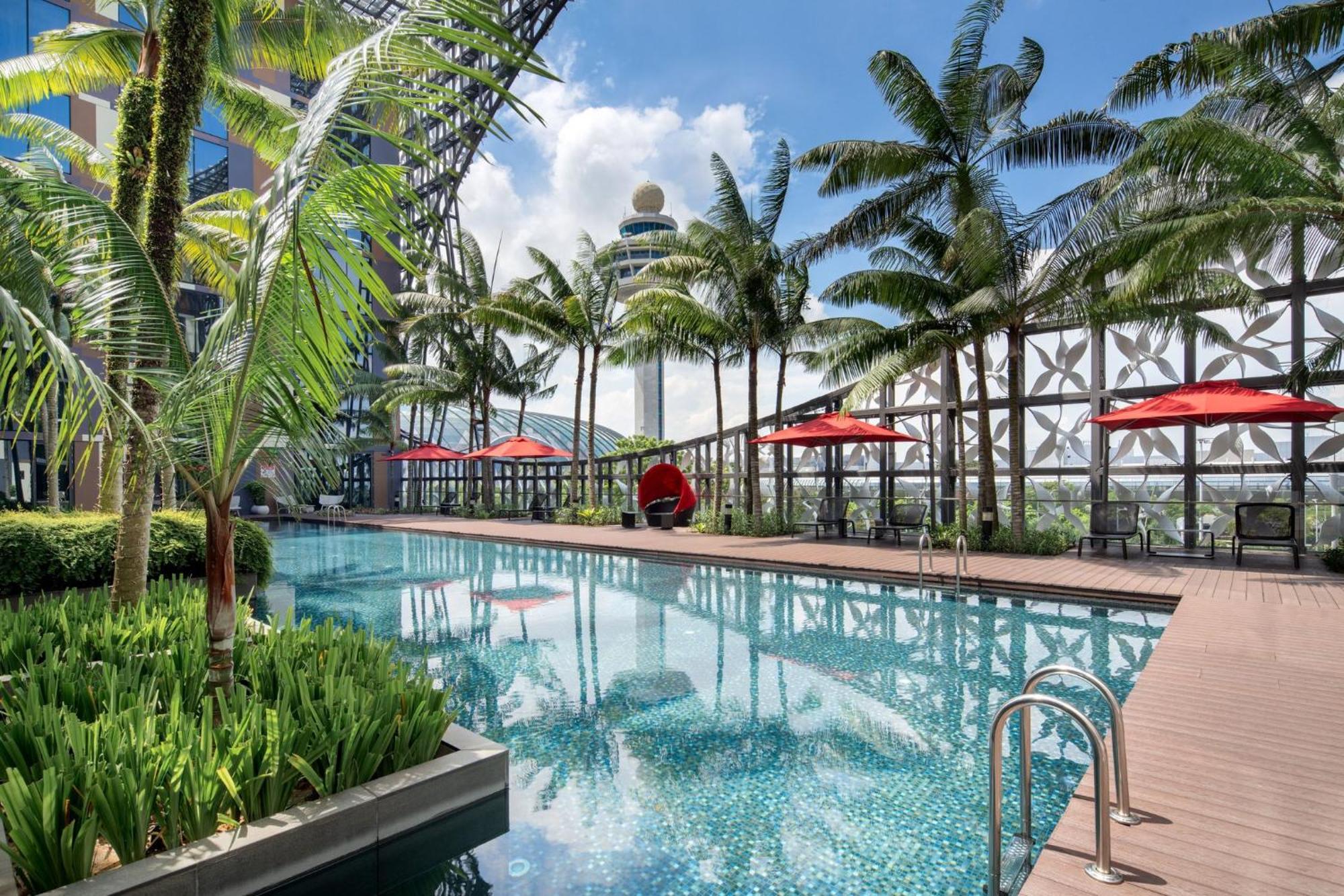 فندق كراون بلازا مطار شانجي سنغافورة المظهر الخارجي الصورة The image shows a luxurious pool area surrounded by palm trees. The pool has a clear blue water surface, with small tiles reflecting light. On one side, there are lounge chairs with red umbrellas providing shade. There are also some decorative plants